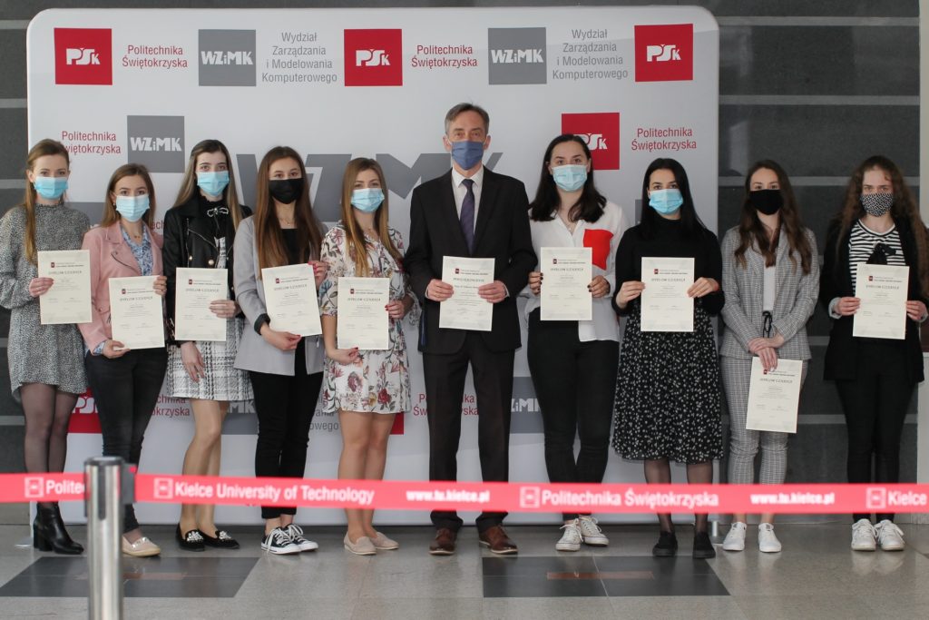 Osoby w maseczkach trzymają dyplomy przed banerem Politechniki Świętokrzyskiej. Kontekst: wręczenie dyplomów.