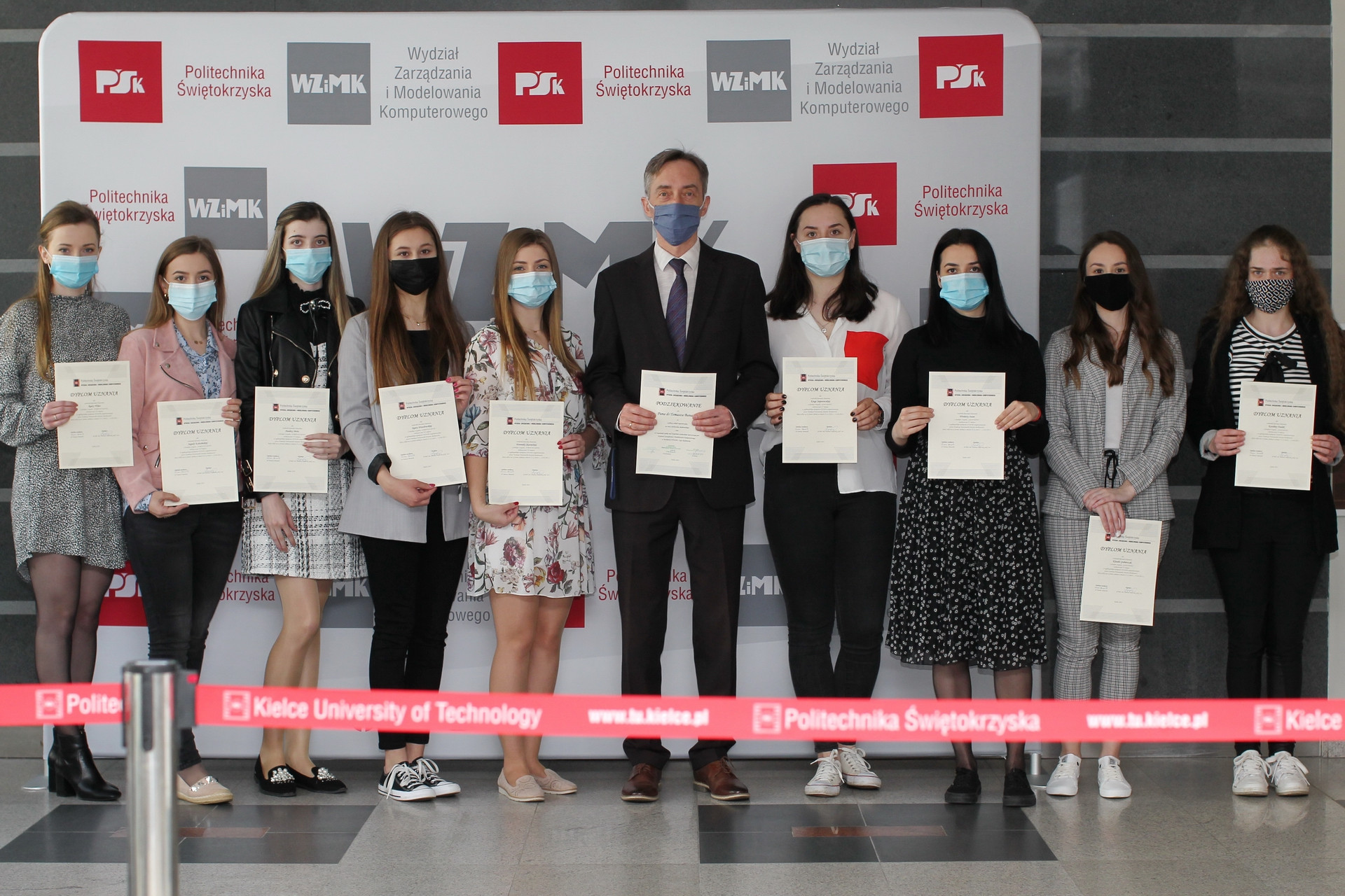 Osoby w maseczkach trzymają dyplomy przed banerem Politechniki Świętokrzyskiej. Kontekst: wręczenie dyplomów.