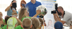 Dzieci w zielonych koszulkach obserwują eksperyment z kulą. Fotograf dokumentuje wydarzenie.