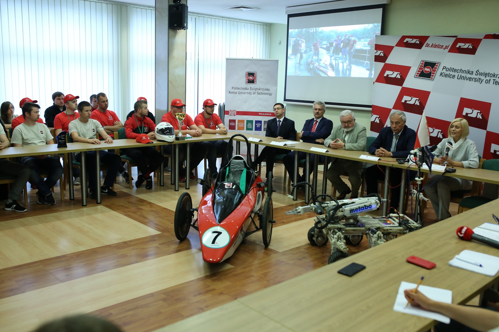 Uczestnicy spotkania przy stole, przed nimi bolid wyścigowy i robot. Kontekst: Politechnika Świętokrzyska.
