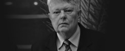 Czarno-białe zdjęcie mężczyzny w garniturze i krawacie. Tło stanowi ciemna roślinność.