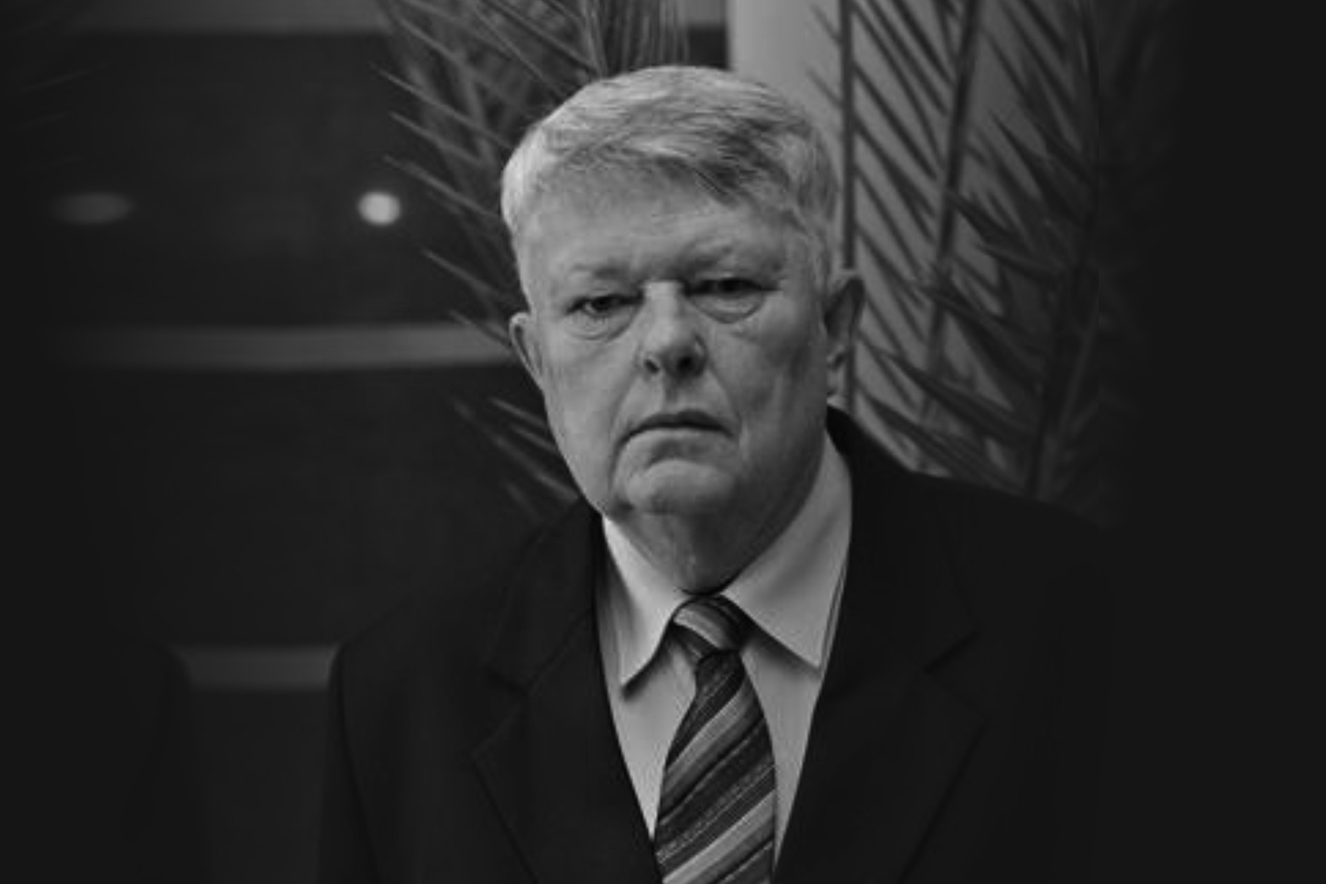 Czarno-białe zdjęcie mężczyzny w garniturze i krawacie. Tło stanowi ciemna roślinność.