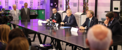 Konferencja prasowa w nowoczesnym laboratorium. Kluczowe obiekty: mężczyźni w garniturach, mikrofony, stoły.