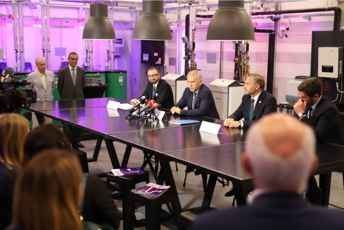Konferencja prasowa w nowoczesnym laboratorium. Kluczowe obiekty: mężczyźni w garniturach, mikrofony, stoły.