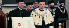 Akademicy w togach i biretach, jeden z nich trzyma dokument. Kontekst: uroczystość akademicka.