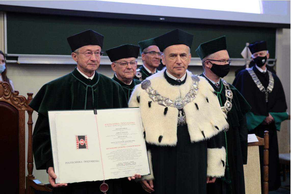 Akademicy w togach i biretach, jeden z nich trzyma dokument. Kontekst: uroczystość akademicka.