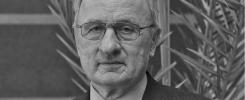 Portret starszego mężczyzny w okularach i garniturze. Tło stanowi roślinność.