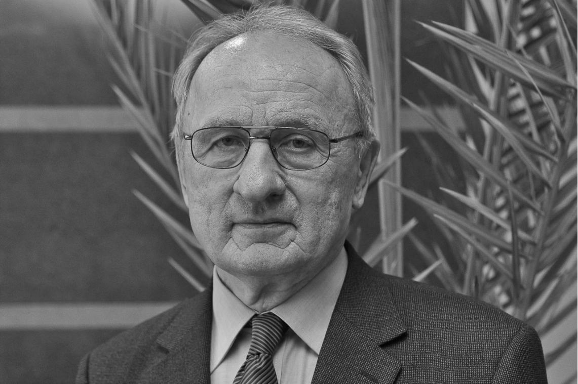 Portret starszego mężczyzny w okularach i garniturze. Tło stanowi roślinność.